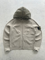 A Stone Island zip-up hooded jacket in a light beige/taupe color, laid flat on a textured gray concrete surface. The iconic black and yellow compass patch is visible on the left sleeve.