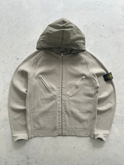 A Stone Island zip-up hooded jacket in a light beige/taupe color, laid flat on a textured gray concrete surface. The iconic black and yellow compass patch is visible on the left sleeve.