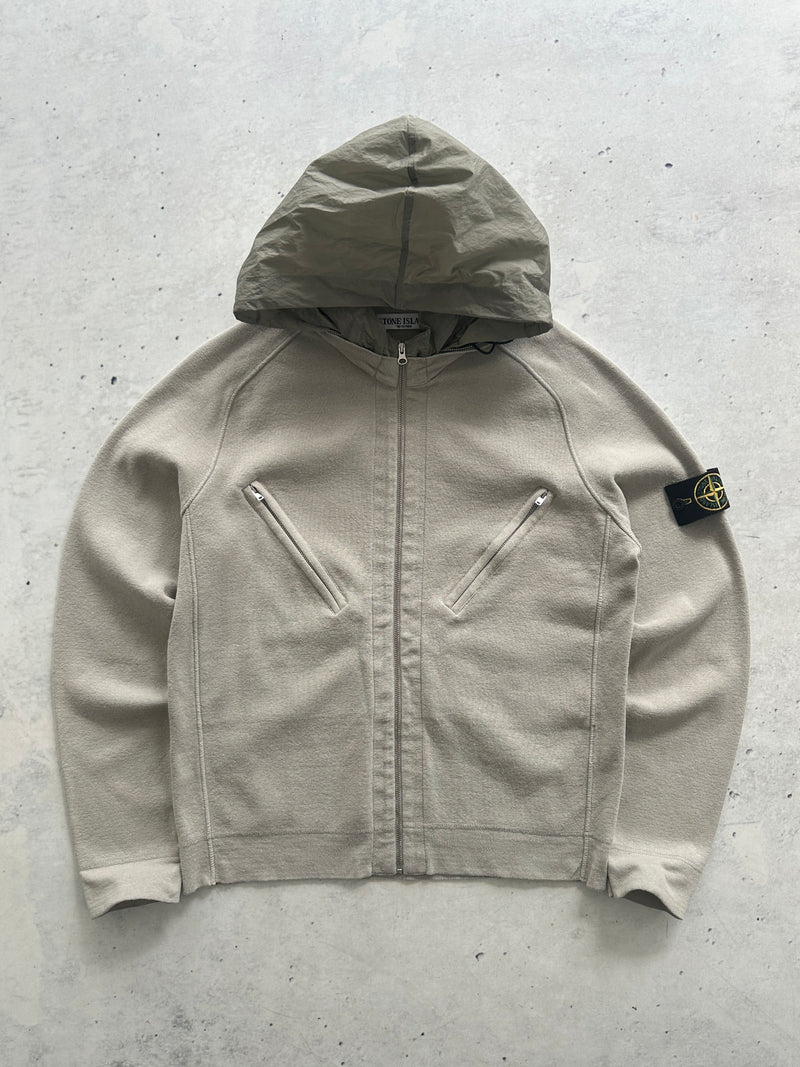 A Stone Island zip-up hooded jacket in a light beige/taupe color, laid flat on a textured gray concrete surface. The iconic black and yellow compass patch is visible on the left sleeve.