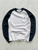 A long-sleeve raglan shirt with a white body and black sleeves, featuring a small embroidered A Bathing Ape logo on the left chest, laid flat on a textured gray concrete surface.