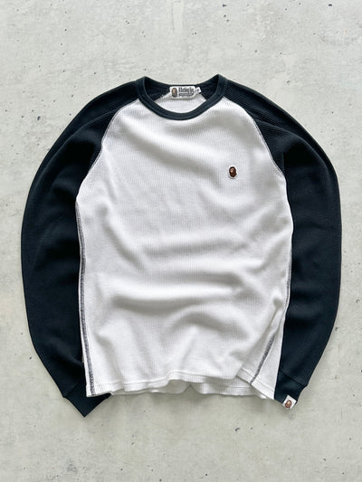 A long-sleeve raglan shirt with a white body and black sleeves, featuring a small embroidered A Bathing Ape logo on the left chest, laid flat on a textured gray concrete surface.