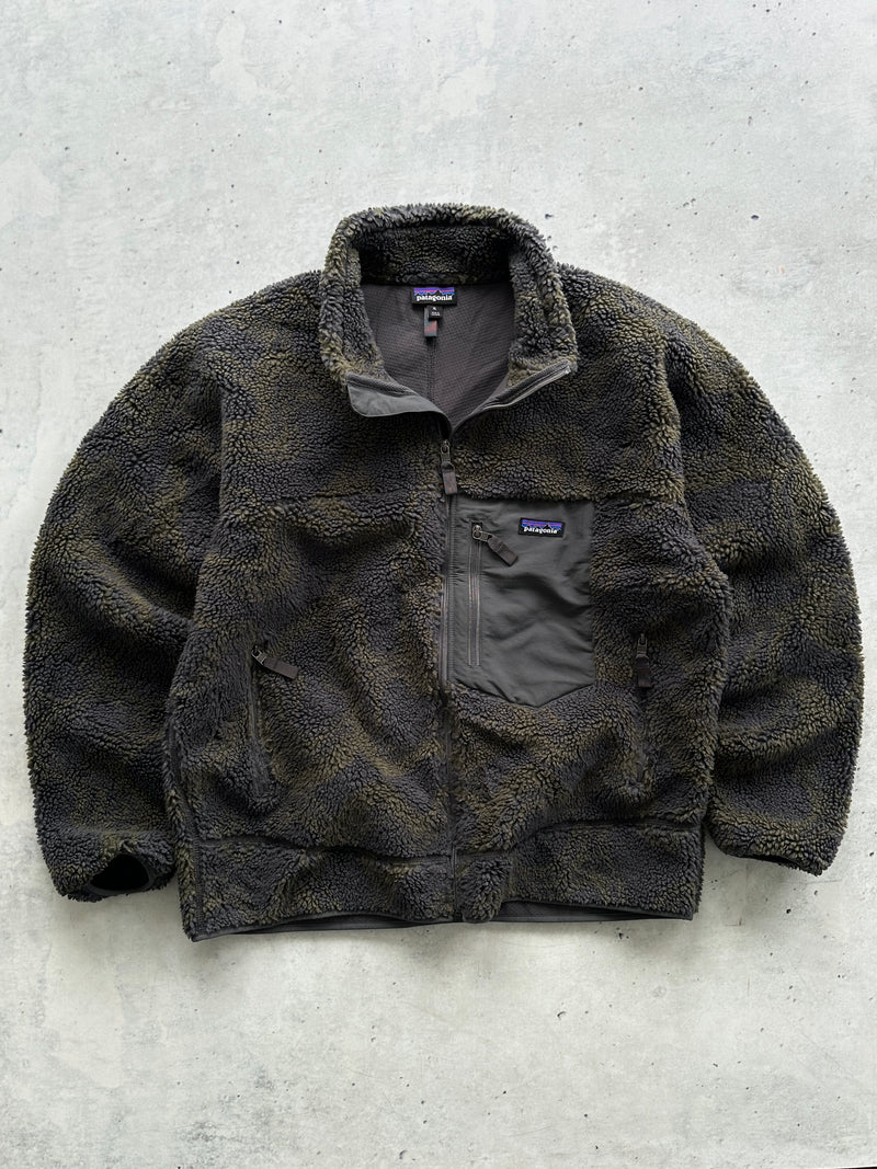 A Patagonia fleece jacket in a dark brown and black textured pattern, laid flat on a light gray concrete surface.