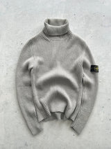 A light gray ribbed knit turtleneck sweater with a Stone Island badge on the sleeve, laid flat on a textured gray concrete surface.