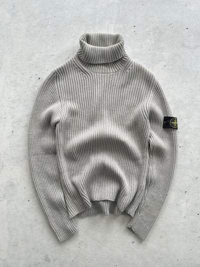 A light gray ribbed knit turtleneck sweater with a Stone Island badge on the sleeve, laid flat on a textured gray concrete surface.