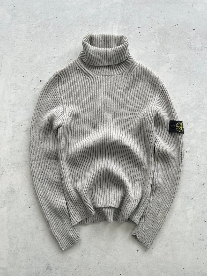 A light gray ribbed knit turtleneck sweater with a Stone Island badge on the sleeve, laid flat on a textured gray concrete surface.