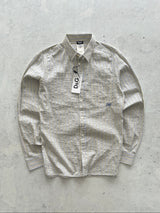 A D&G long-sleeve button-up shirt in a beige and white textured stripe pattern, laid flat on a light gray concrete surface with a paper tag still attached.