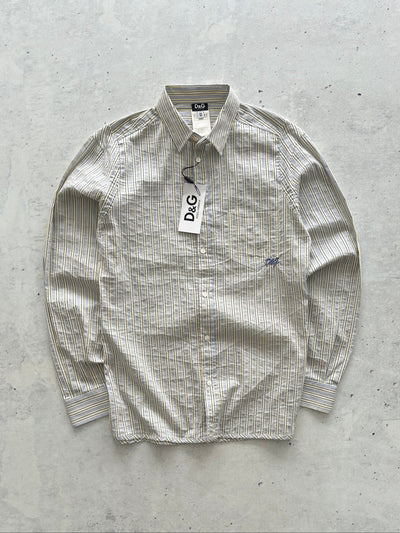 A D&G long-sleeve button-up shirt in a beige and white textured stripe pattern, laid flat on a light gray concrete surface with a paper tag still attached.