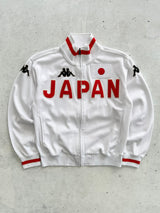 A white Kappa track jacket featuring 'JAPAN' printed in bold red letters across the chest, a Japanese flag patch on the left chest, and red and black trim on the collar and cuffs, laid flat on a textured gray background.