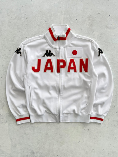 A white Kappa track jacket featuring 'JAPAN' printed in bold red letters across the chest, a Japanese flag patch on the left chest, and red and black trim on the collar and cuffs, laid flat on a textured gray background.