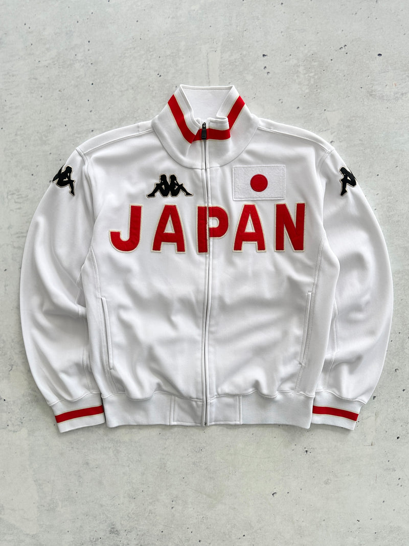 A white Kappa track jacket featuring 'JAPAN' printed in bold red letters across the chest, a Japanese flag patch on the left chest, and red and black trim on the collar and cuffs, laid flat on a textured gray background.