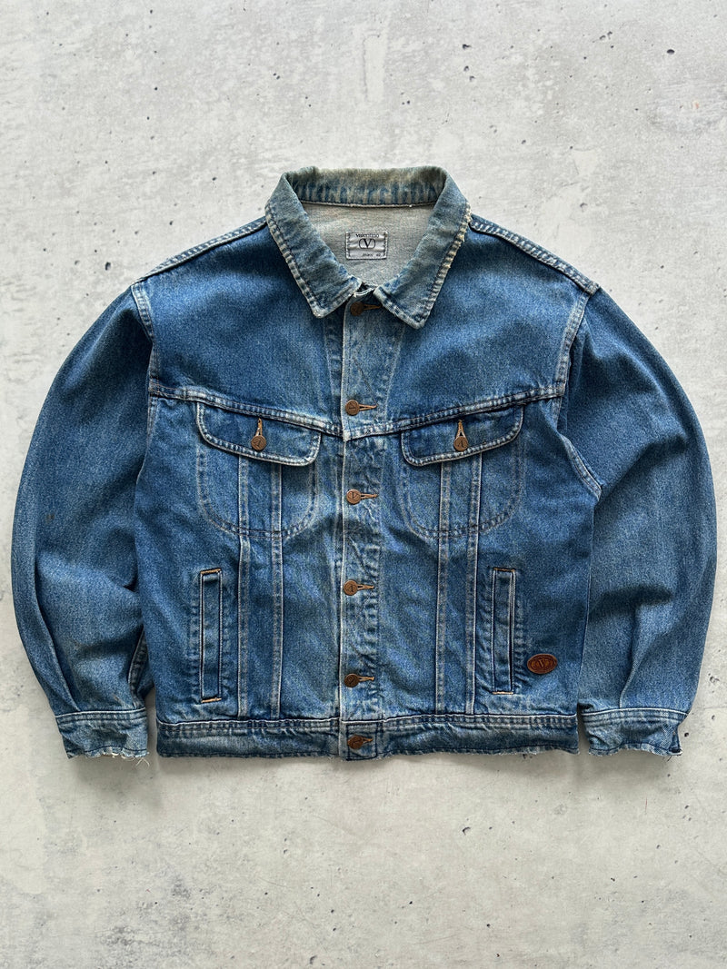 A classic blue Valentino vintage denim jacket with a button-up front, chest flap pockets, and side slip pockets, laid flat on a textured gray concrete surface.